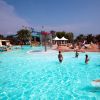 Piscina all'aperto con persone che nuotano, scivoli sullo sfondo e palme attorno.