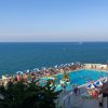 Piscina affollata vicino al mare, ombrelloni blu e bianchi, persone che nuotano e prendono il sole.