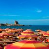 Spiaggia sabbiosa con ombrelloni arancioni e gialli, mare azzurro e torre costiera sullo sfondo.