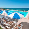 Piscina all'aperto con ombrelloni blu e bianchi vicino al mare, persone rilassate sui lettini.