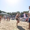 Persone che giocano in spiaggia, un uomo tiene una maglietta mentre due ragazze abbracciate saltano.