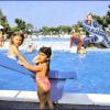Bambine accanto a una piscina con scivolo, altre persone in acqua.